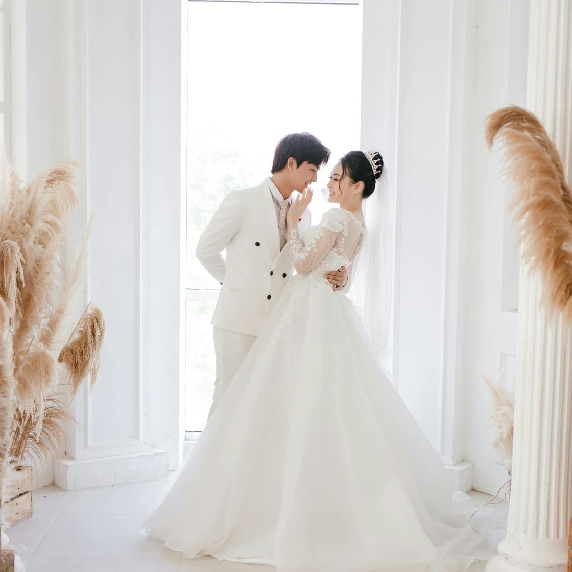 Asian couple dressed in white, standing in a bright room with white walls, about to kiss near a tall door with abundant light coming through
